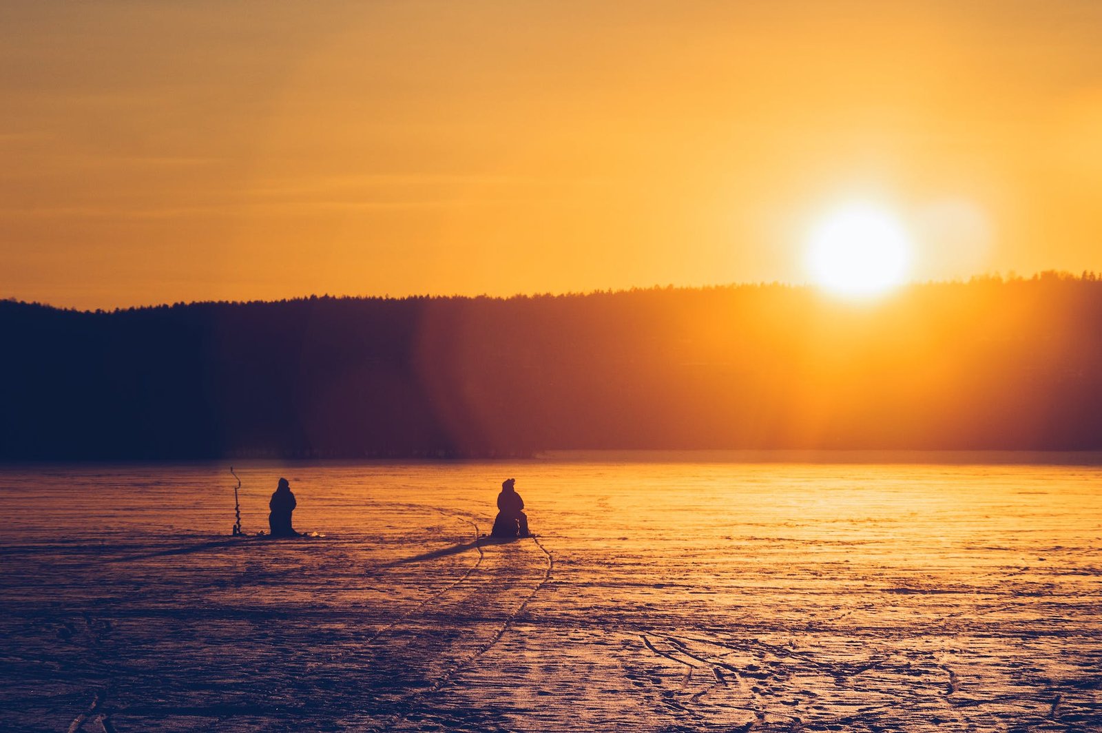 silhouette of two persons sitting while snow fishing on an iced covered body of water at dawn - hiking in finland